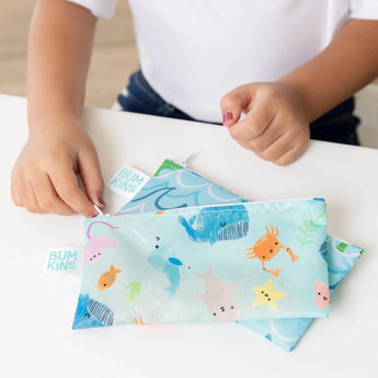 A child holds a Bumkins snack bag with ocean life designs, including crab and fish, on a white table.