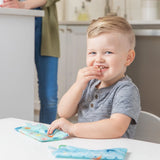 A young child smiles while snacking from a Bumkins Reusable Snack Bag, Ocean Life design. An adult stands in the kitchen behind them.