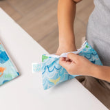 Child with a Bumkins Ocean Life Snack Bag, part of a small 2-pack, holding it at a white table.