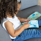 A curly-haired child, in a white top and blue jeans, holds a Bumkins Ocean Life reusable snack bag on the couch.