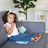 Toddler with curly hair and a bow enjoys snacks from Bumkins Reusable Snack Bags on a gray sofa, wearing a white top and jeans.