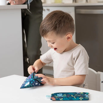 A child eagerly unpacks a Bumkins Reusable Snack Bag, Small 2-Pack: Jungle Prints, at the table; an Animal Print pouch is nearby.