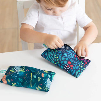 A child in a white shirt examines the jungle and animal print waterproof fabric of Bumkins Reusable Snack Bag, Small 2-Pack at a table.