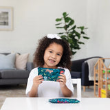 A curly-haired child holds a colorful pencil case and Bumkins reusable snack bags, sitting at a table in a cozy room with toys.