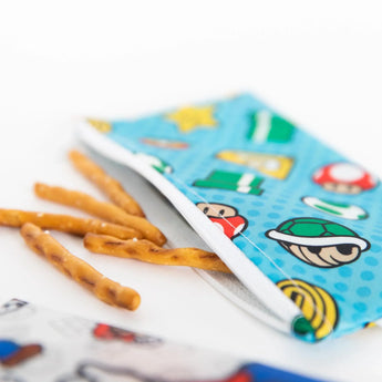 Pretzel sticks spill from Bumkins waterproof Reusable Snack Bag in the Super Mario™ Small 2-Pack, partially open on a white surface.