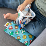 A child sits barefoot on a gray surface with Bumkins Reusable Snack Bag, Small 2-Pack: Super Mario™, one open and ready for fun snacks.