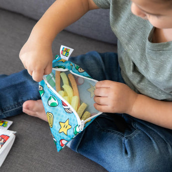 Child on couch reaching into colorful, waterproof Bumkins Reusable Snack Bag, Small 2-Pack: Super Mario™ for candy strips.