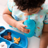 A toddler develops drinking skills using a Bumkins Silicone Straw Cup with Lid: Blue, plus a colorful bib and LFGB-grade silicone tray.