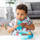 A toddler with a Mickey Mouse bib masters drinking independently using the Bumkins Silicone Straw Cup with Lid: Blue at the table.