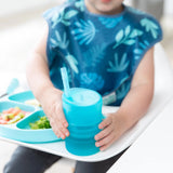 A toddler in a blue bib holds a Bumkins Silicone Straw Cup with Lid: Blue, sitting at a highchair beside a plate of varied foods.