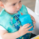 A toddler uses a Bumkins Silicone Straw Cup with Lid in blue, drinking independently indoors while wearing a bib.