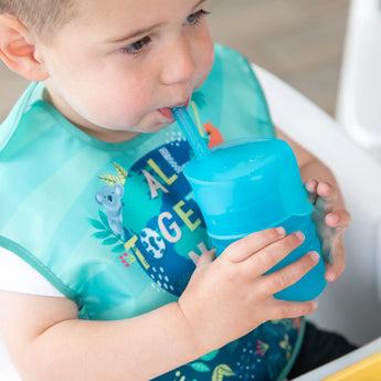 A toddler uses a Bumkins Silicone Straw Cup with Lid in blue, drinking independently indoors while wearing a bib.