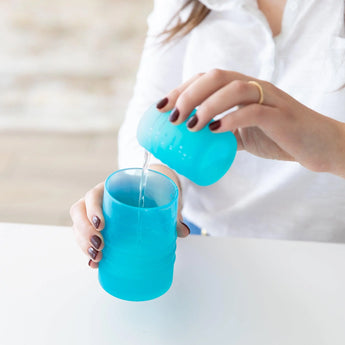 Someone pours liquid from a small LFGB-grade silicone cup into a larger Bumkins Silicone Straw Cup with Lid: Blue.