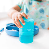 A toddler holds a Bumkins Silicone Straw Cup with Lid: Blue, while an LFGB-grade silicone plate sits on the table in the background.