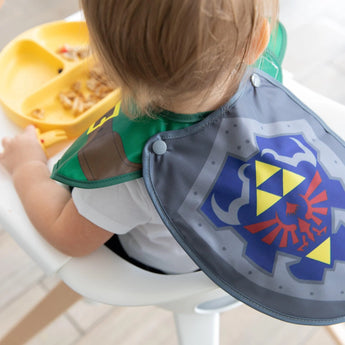 A child in a high chair wears a Bumkins Caped SuperBib®: The Legend of Zelda™ Link, near a yellow plate with food scraps.