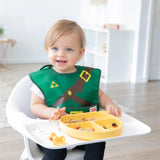 A baby in a highchair wears a Bumkins Caped SuperBib® Zelda Link while dining on a divided yellow plate of food on the tray.