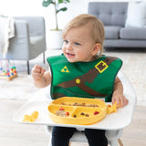 A baby in a high chair wears a Bumkins Caped SuperBib®: The Legend of Zelda™ Link, holding food as a divided plate awaits.