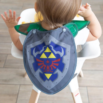 A toddler in a high chair eagerly reaches out wearing a Bumkins Caped SuperBib®: The Legend of Zelda™ Link, with easy-wipe shield design.