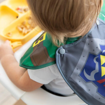 A child in a high chair wears Bumkins Caped SuperBib®: The Legend of Zelda™ Link, ready to eat with the easy-wipe bib for protection.