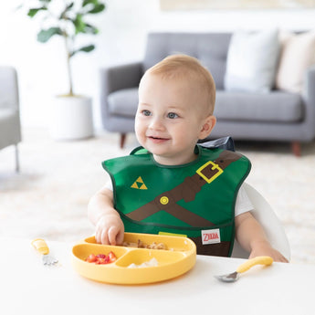 A baby in a green Bumkins Caped SuperBib®: The Legend of Zelda™ Link sits at a table with a yellow divided plate and cutlery.