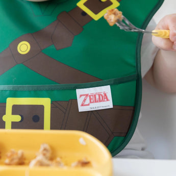 A child in the Bumkins Caped SuperBib®: The Legend of Zelda™ Link enjoys food from a yellow tray, keeping mealtime mess-free.