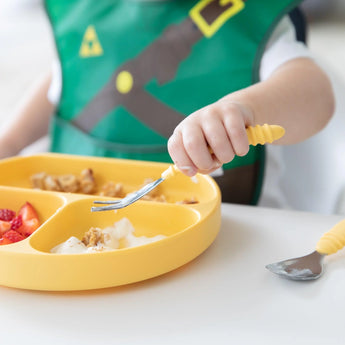 A child in a green Caped SuperBib® enjoys strawberries, yogurt, and cereal from a yellow plate with a fork labeled Bumkins.