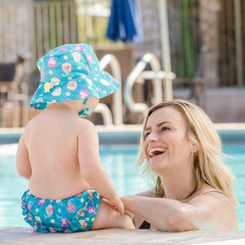 A smiling woman in a pool interacts with a baby wearing the Bumkins Swim Set: Mermaids with UPF 50+ sun hat at the pools edge.