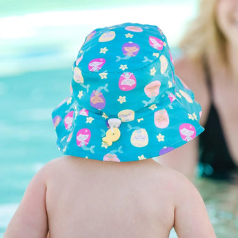 A baby in a Bumkins Swim Set: Mermaids with a sun hat and swim diaper is poolside, watched by an adult nearby.