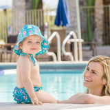 A baby in a Bumkins Swim Set: Mermaids sits by the pool, while a woman in the water smiles at them.