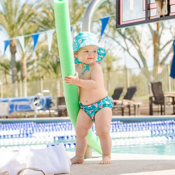 A baby in a Bumkins Mermaids set holds a green noodle, backed by palm trees and lounge chairs.