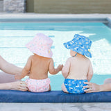 Two babies sit by a pool with adults hands supporting them, wearing Bumkins Swim Diapers, one pink and one blue.