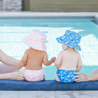 Two babies sit by a pool with adults hands supporting them, wearing Bumkins Swim Diapers, one pink and one blue.