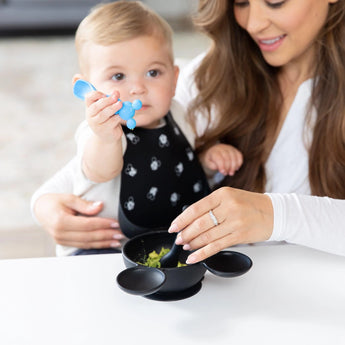 A baby with a Mickey Mouse bib holds a Bumkins Silicone Dipping Spoon as a woman helps mix food in the black bowl.