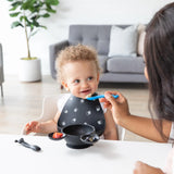 A smiling toddler in a black bib is fed with Bumkins blue Mickey Mouse spoon; a bowl and silicone spoons are ready for self-feeding.