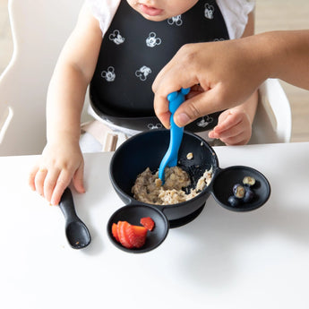 A baby in a high chair with a bib featuring mouse ears self-feeds strawberries and blueberries using Bumkins Mickey Mouse silicone spoons.