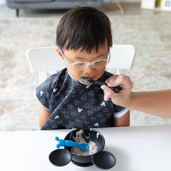 A child happily enjoys yogurt with a Bumkins Mickey Mouse Silicone Dipping Spoon at the table.