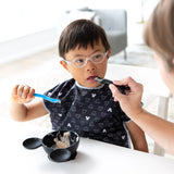 A child with glasses self-feeds using a blue fork, assisted by an adult with a Bumkins Mickey Mouse bowl and silicone spoon.