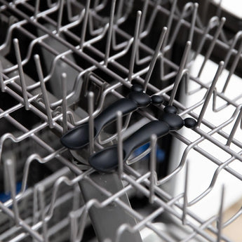 On the top rack of an open dishwasher, pliers rest among gray metal spikes and Bumkins Mickey Mouse Silicone Dipping Spoons.