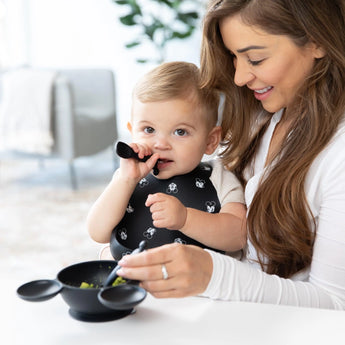 A woman smiles as the baby uses a Bumkins Mickey Mouse Silicone Dipping Spoon and bib, exploring self-feeding near a handled bowl.