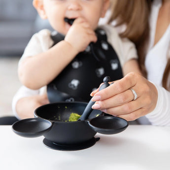 A baby in a black bib is fed green food with a Bumkins Mickey Mouse Silicone Dipping Spoon from a food-safe silicone bowl.