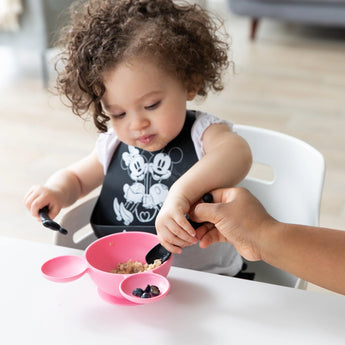 A toddler enjoys a meal with a Mickey Mouse bib and Bumkins silicone dipping spoon, assisted by an adults hand, using a pink bowl.