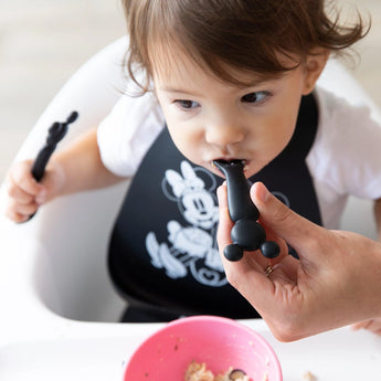 Toddler with a bib using Bumkins Mickey Mouse Silicone Dipping Spoons while holding a matching fork over a pink bowl on their high chair.