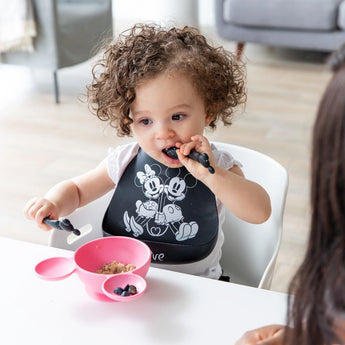 A curly-haired toddler enjoys blueberries from a pink bowl, using black Mickey Mouse spoons, while wearing a matching bib at the table.