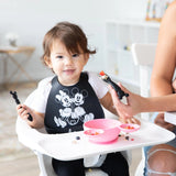 A toddler in a bib reaches for Bumkins Mickey Mouse Silicone Dipping Spoons as bites are offered from a pink bowl.