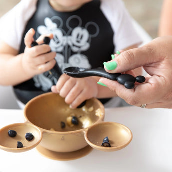 A child enjoys blueberries using Bumkins food-safe silicone Mickey Mouse spoons and bowl, learning to self-feed with adult help.