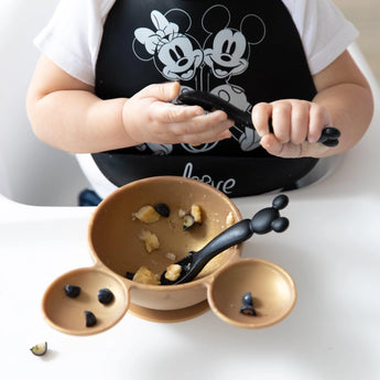 A child in a Mickey Mouse bib uses Bumkins food-safe Silicone Dipping Spoons with a divided brown bowl on a highchair tray.