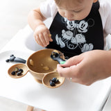 A toddler in a high chair eats blueberries from a Mickey Mouse bowl using Bumkins Silicone Dipping Spoons, wearing a Mickey and Minnie bib.