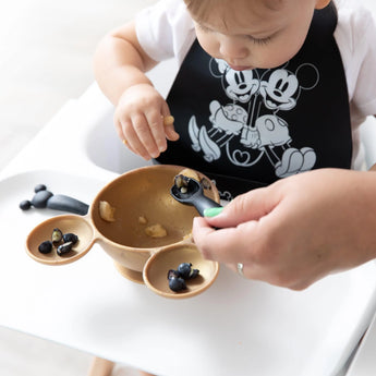 A toddler in a high chair eats blueberries from a Mickey Mouse bowl using Bumkins Silicone Dipping Spoons, wearing a Mickey and Minnie bib.