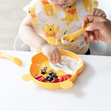 A baby in a bib explores self-feeding with Bumkins Winnie the Pooh silicone spoons, reaching for fruit on a yellow bear plate, aided by yogurt.