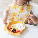 A child enjoys yogurt with fruit on a bear plate, using Bumkins Winnie the Pooh Silicone Dipping Spoons with adult assistance.